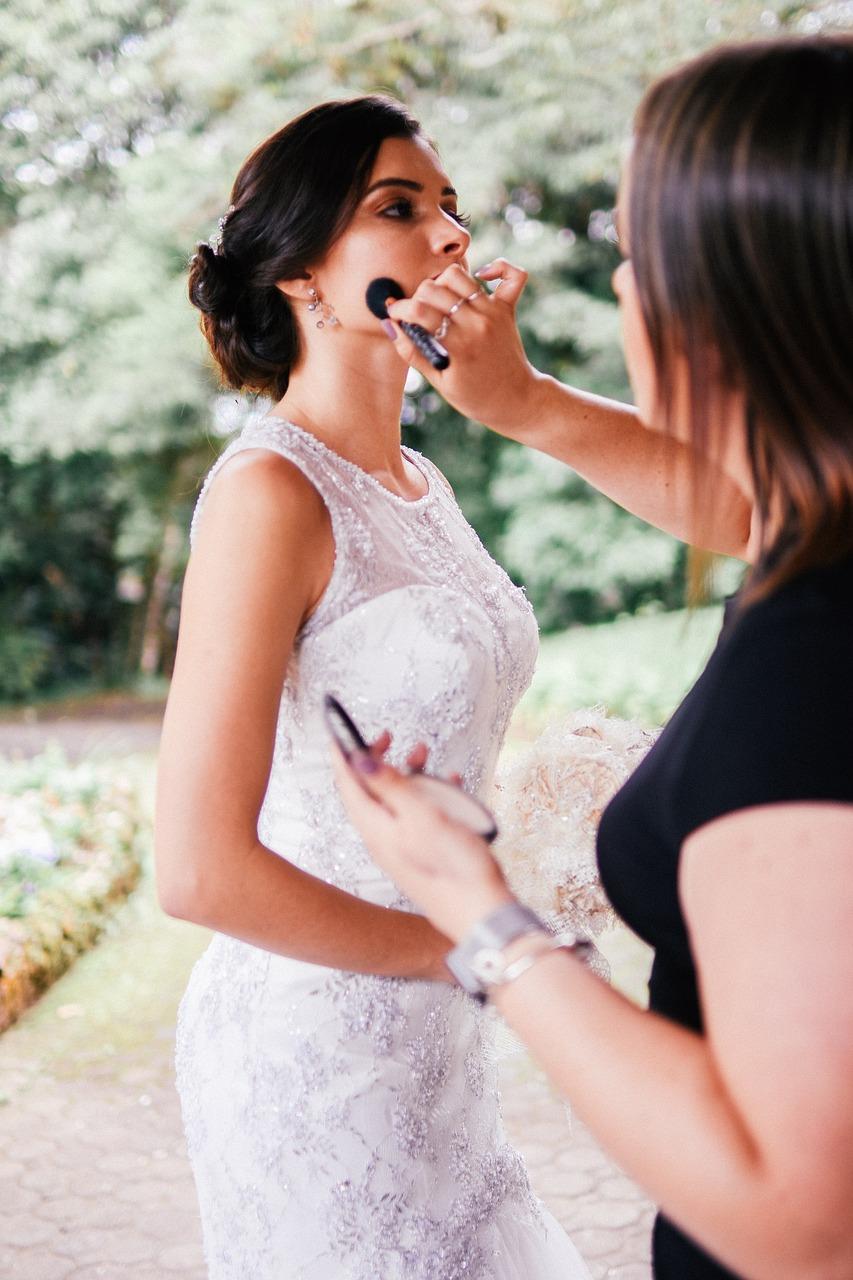 Makeup artist applying blush to a bride wearing an intricate lace wedding dress during outdoor wedding preparations