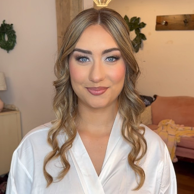 Bride wearing white lace-trimmed robe with romantic loose curls, glam makeup, and rosy cheeks in bridal suite