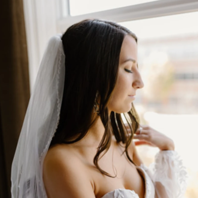 Elegant bride in off-shoulder lace gown and cathedral veil, with dark hair styled straight and romantic natural makeup, gazing by window